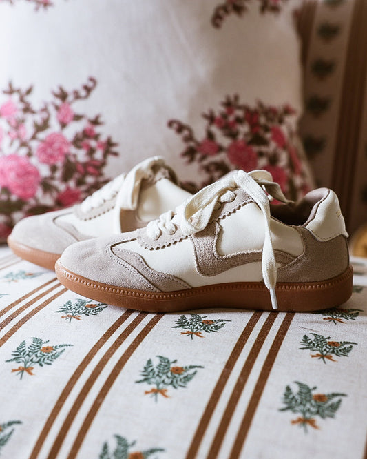 A pair of beige and off-white low-top sneakers with brown soles, resting on a patterned sofa cushion with floral pillows in the background.