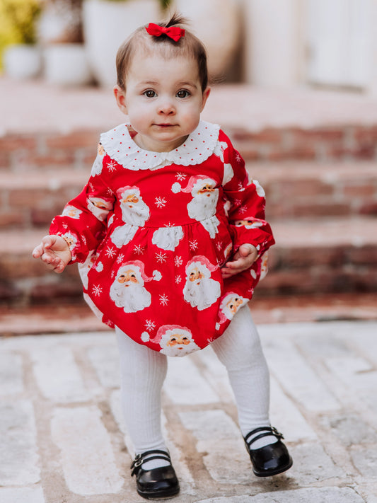 Baby girl outside wearing a jolly santa print classic bubble with white tights and black mary janes.