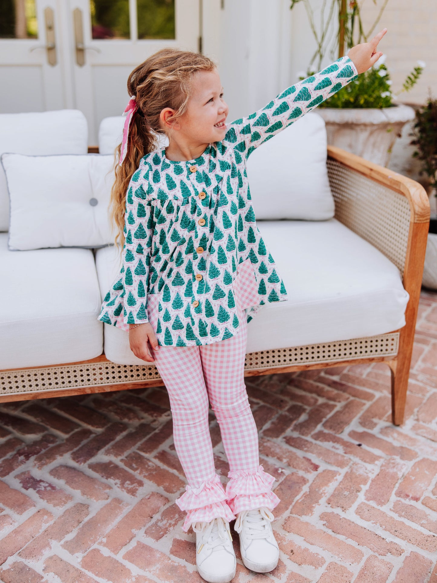 Girl holding arms out wearing a Christmas tree themed out to play set with pink gingham bottoms and white tennis shoes.