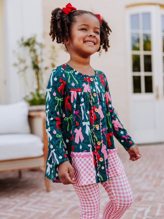Girl wearing a holiday themed two piece set. Top has colorful bows and bottoms and accent pockets are pink gingham.