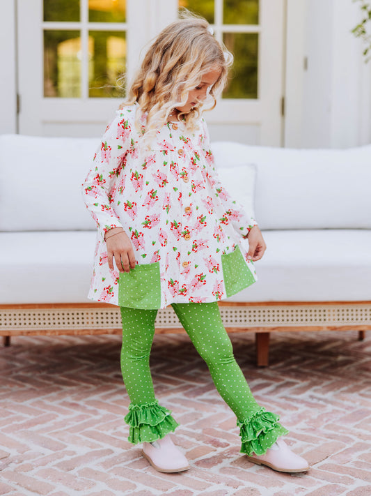 Girl standing outside in front of couch wearing a holiday themed top with green leggings with white polka dots that have ruffles at bottom.