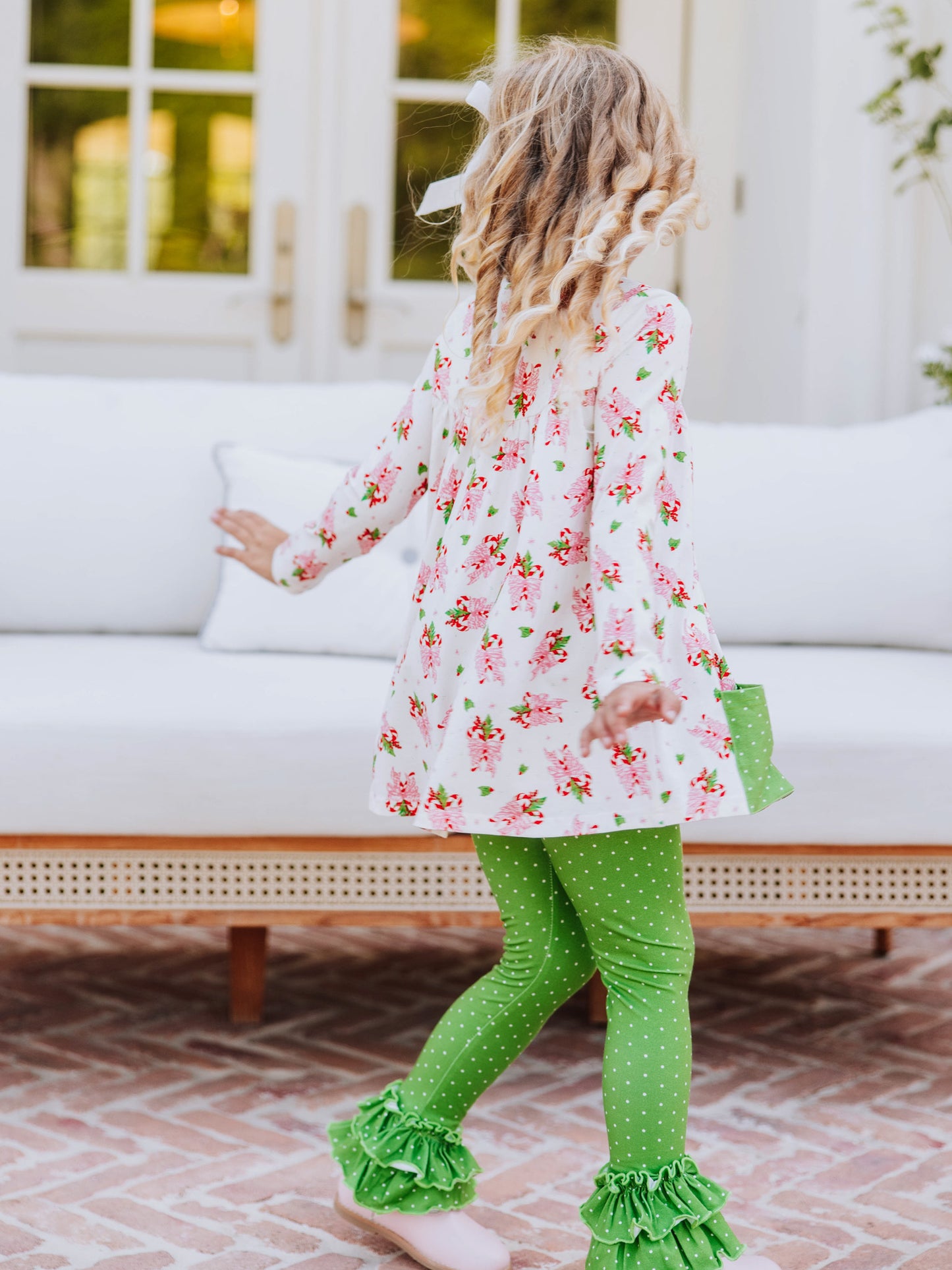 Girl standing outside in front of couch wearing a holiday themed top with green leggings with white polka dots that have ruffles at bottom.