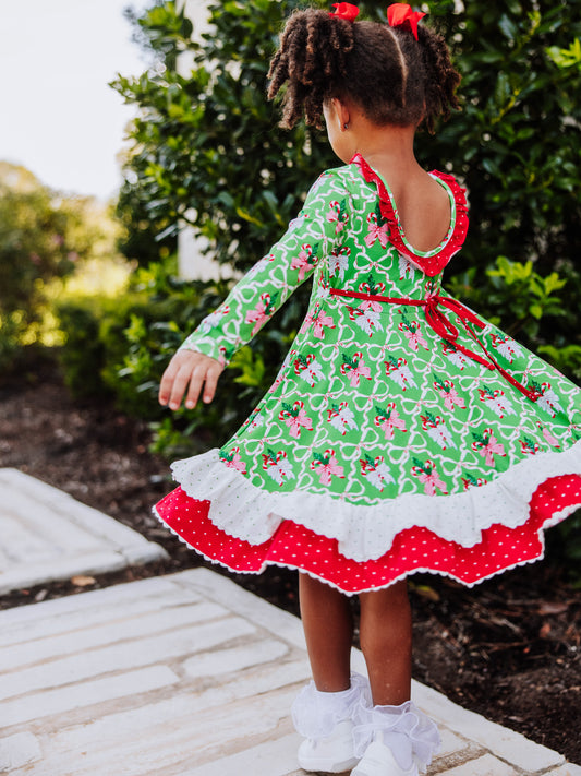 Girl is standing outside holding arms out. She is wearing a holiday themed dress with ruffly socks and white shoes. Girl is slowly twirling to show movement in dress.