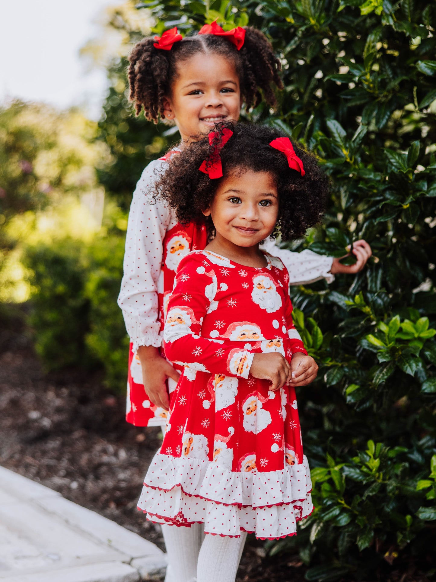 Two models standing near bush smiling at camera. Littlest one wearing a red jolly santa dress.