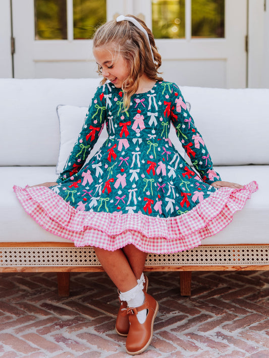 Girl sitting on couch wearing a holiday colorful bow dress that has a double ruffle pink gingham pattern.