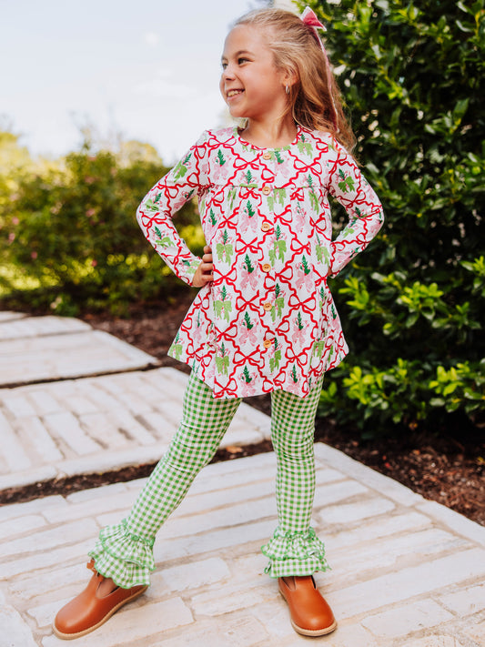 Girl has hands on her hips and is wearing a holiday bow themed top with green gingham bottoms with ruffles at ankles.