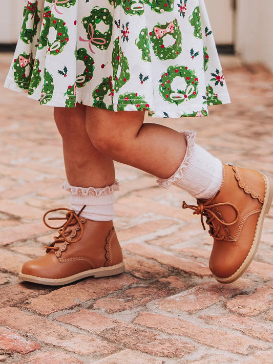 Girl outside standing on brick wearing a pair of lacy ankle boots with scalloped detail on them. Model is also wearing pink socks and a dress with green wreaths on it.