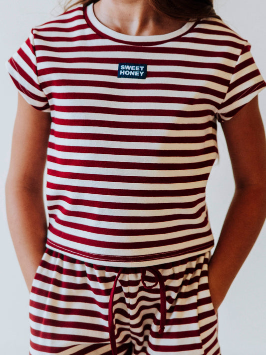 Girl is wearing a two-piece red and ivory striped pant set. Top has a SweetHoney logo in white and navy in the center of chest. Girls hands are in her pocket.