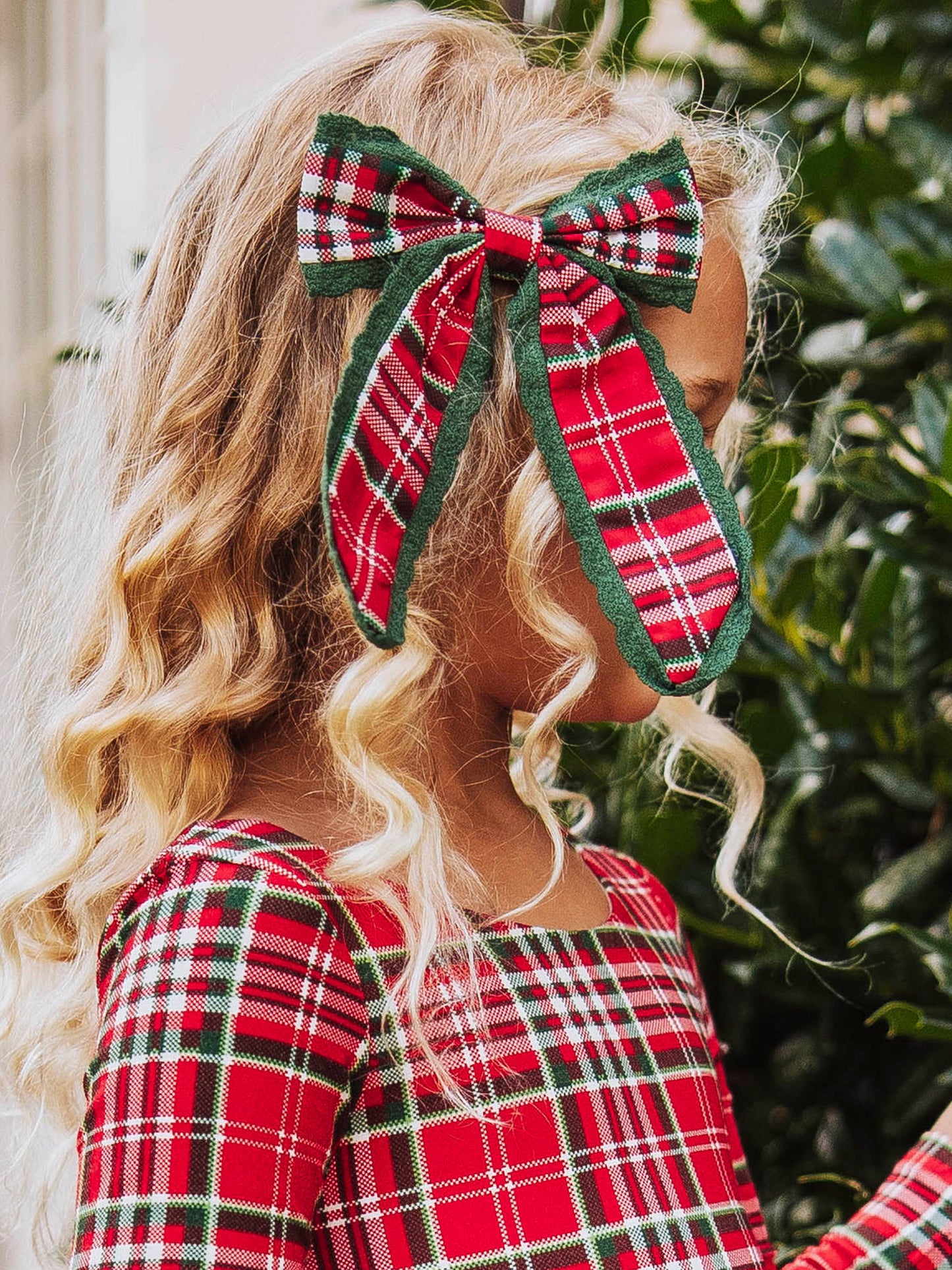Up close shot of girl wearing a red traditional colored plaid bow on the side of her head. Model wearing the same colored leggy romper too.