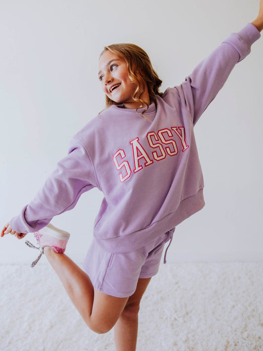 Model is wearing a two piece, purple in color sweat set. the top has SASSY on the front in white letters that are outlined in pink.