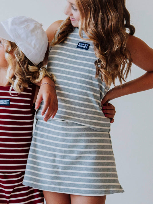 Model is wearing a two-piece gray and white striped skort set. There is a SweetHoney tag that is centered on front of shirt and is navy and white in color. Model is also wearing a white hat.