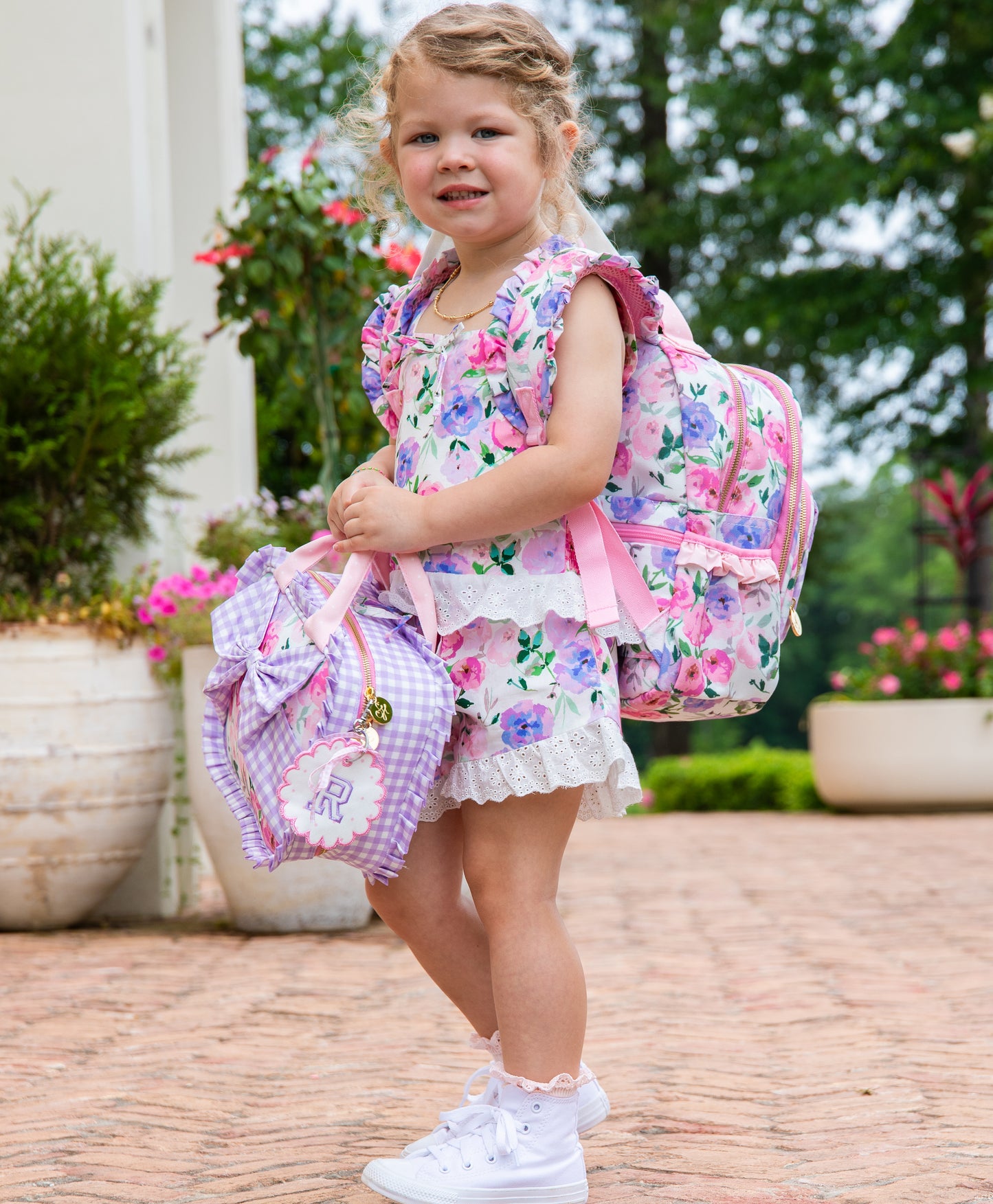 Girl wearing a colorful floral backpack with a purple gingham bow attached to the backpack. Pack also has custom embroidery. Girl also holding same printed style lunch kit