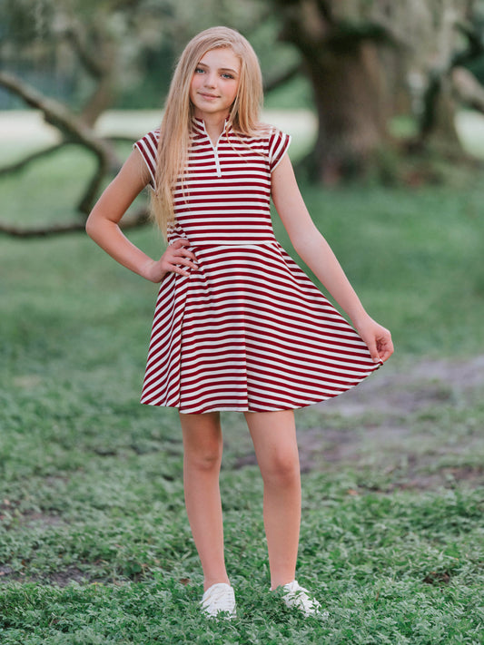 Model outside in wooded green area wearing a crimson and ivory striped dress with white canvas shoes. Model is slightly holding out the side of the dress.