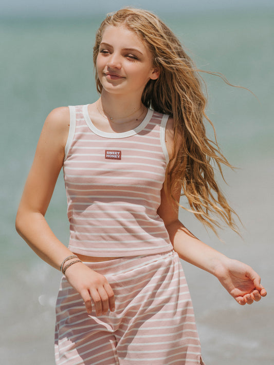 Model is outside on a beach wearing a two-piece matching pink and ivory striped skort set. Top is sleeveless and has a SweetHoney logo tag in the middle of the chest.