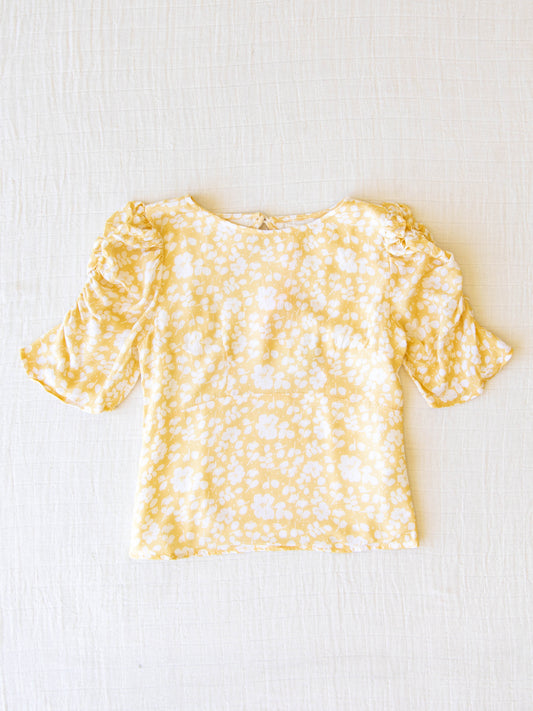 Classic Smocked Cutout Top – I’ve Got Sunshine. This short sleeve top has a smocked and keyhole back, and synched sleeves. It is a pattern of white flowers on a bright yellow background.