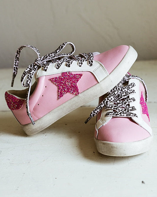 A pair of pink and white low-top sneakers with glittery silver laces and a pink glitter star on the side, resting on a white wooden surface against a beige wall.