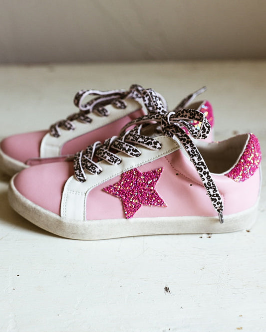 A pair of pink low-top sneakers with white rubber toe caps and soles, decorated with glittery pink star patches on the sides and glittery silver laces, resting on a white wooden surface.
