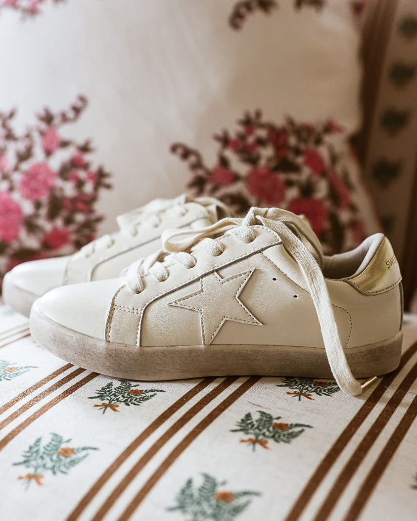 A pair of off-white leather sneakers with star details on the side, resting on a patterned sofa cushion with floral and striped pillows in the background.