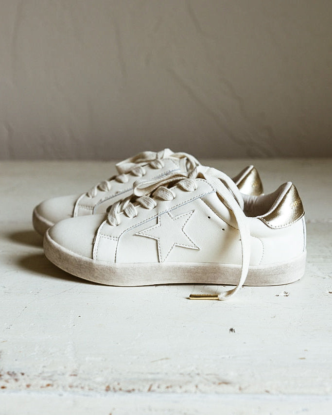 A pair of off-white low-top sneakers with tonal laces and subtle gold-toned heel accents, resting on a distressed white wooden surface against a neutral beige wall.