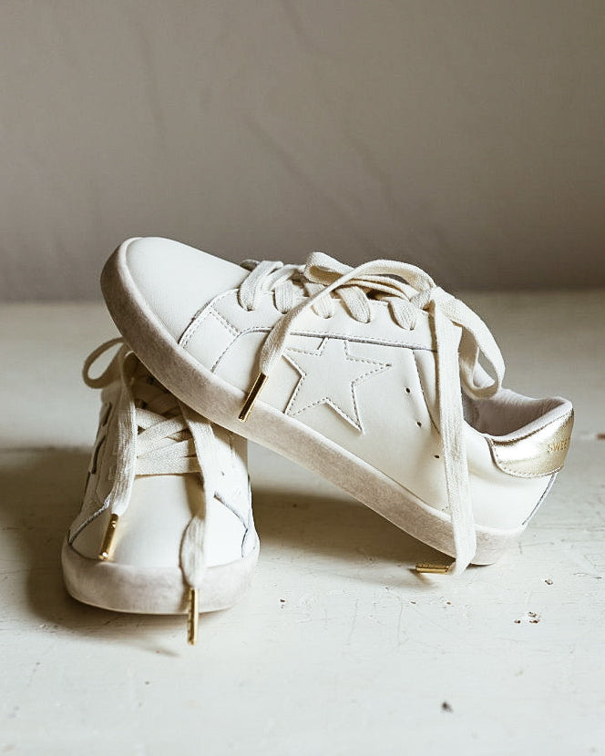 A pair of off-white leather low-top sneakers with tonal star cutouts on the sides, displayed on a rustic white wooden surface against a neutral beige wall.