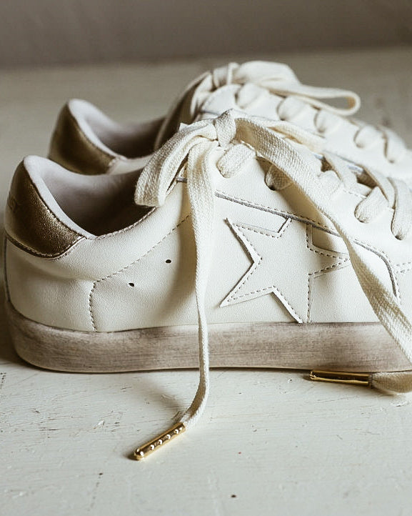 A pair of off-white low-top sneakers with tonal laces and subtle gold-toned heel accents, resting on a distressed white wooden surface against a neutral beige wall.