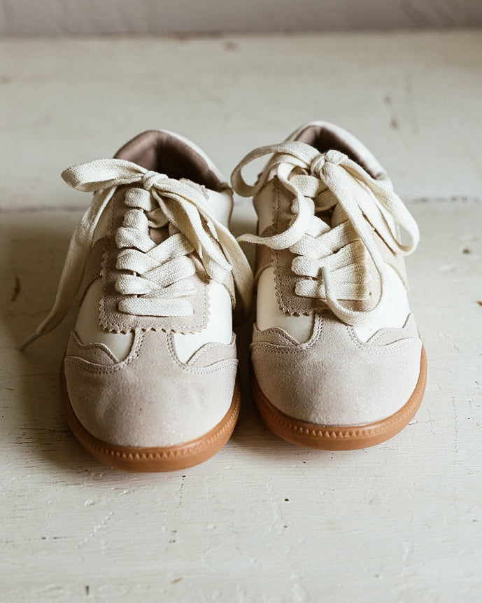 A pair of cream-colored suede sneakers with tan rubber soles, photographed on a rustic, light-colored wooden floor against a neutral wall.