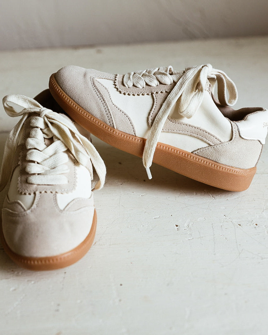 A pair of low-top canvas sneakers in off-white and beige with tan soles, arranged on a light-colored wooden floor.