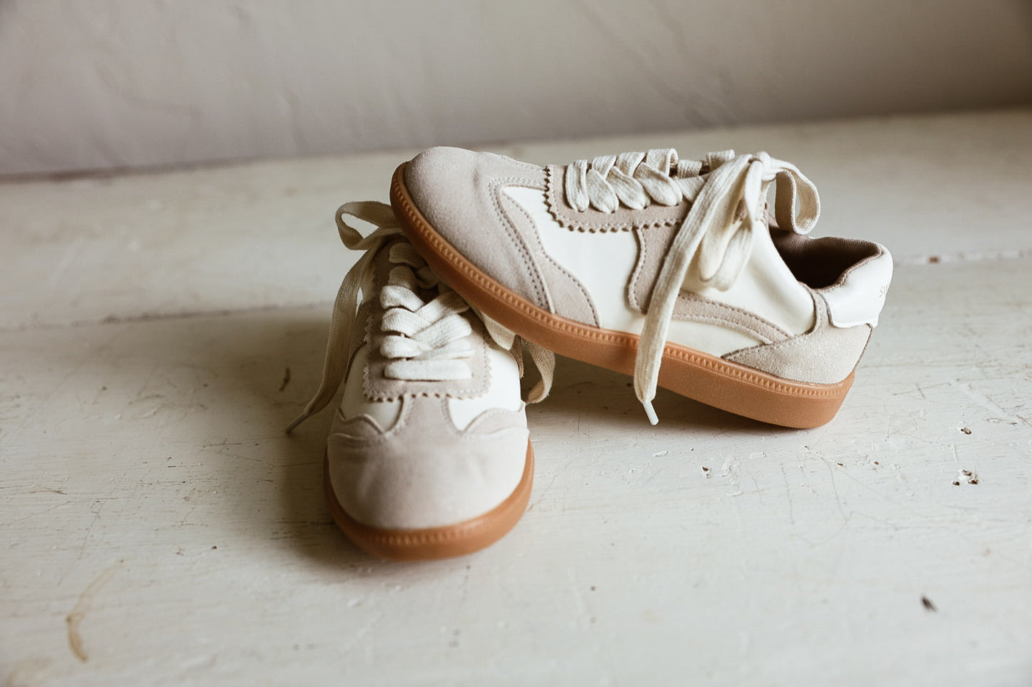 A pair of cream-colored suede sneakers with tan rubber soles, photographed on a rustic, light-colored wooden floor against a neutral wall.