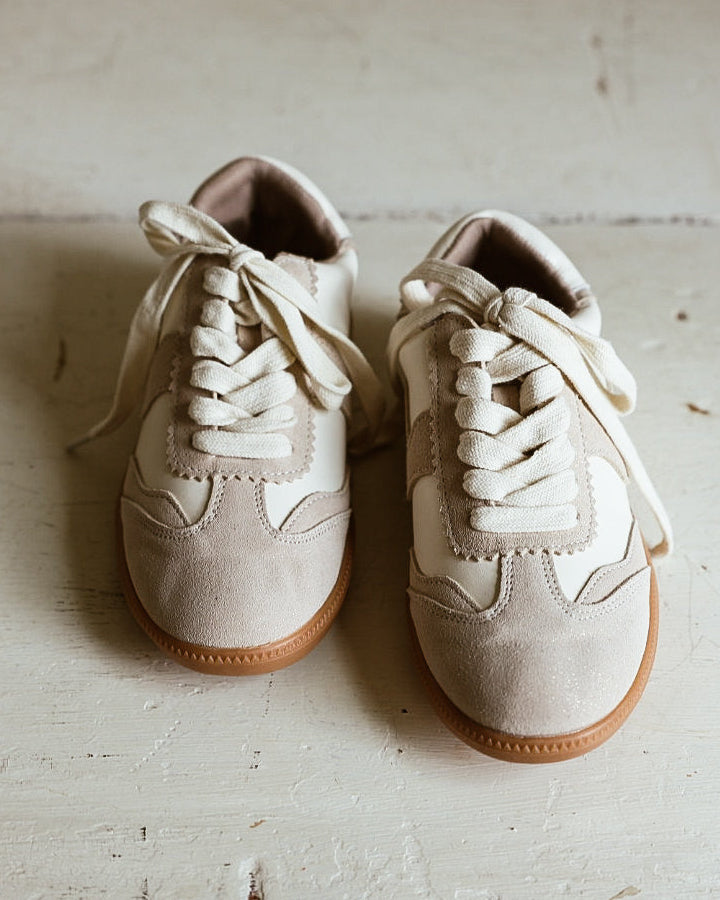 A pair of low-top beige sneakers with white laces and gum soles, placed on a light-colored wooden floor.