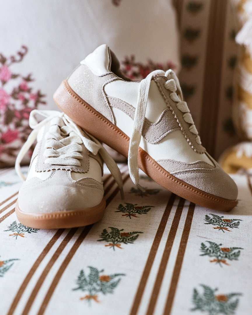 A pair of beige and off-white low-top sneakers with brown soles, resting on a patterned sofa cushion with floral pillows in the background.