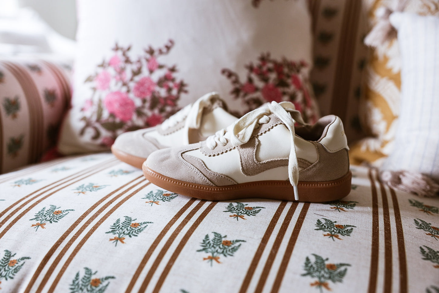A pair of cream-colored suede sneakers with tan rubber soles, photographed on a rustic, light-colored wooden floor against a neutral wall.