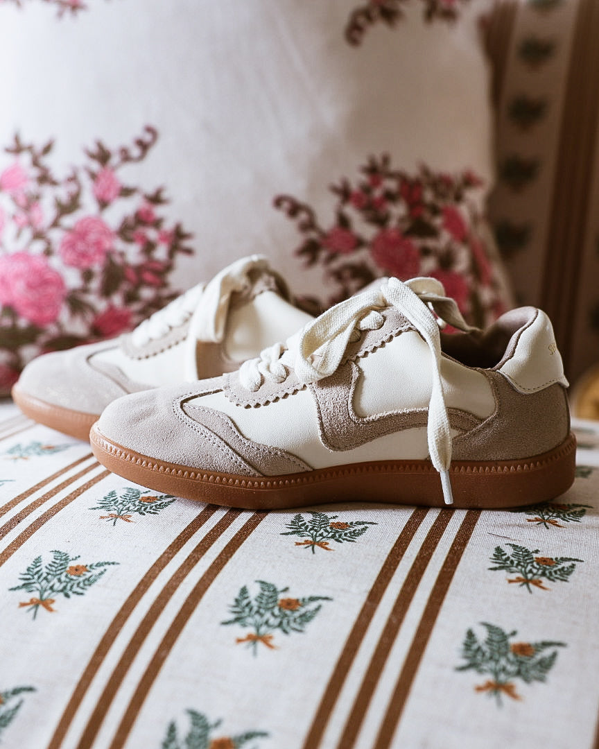 A pair of beige and off-white low-top sneakers with brown soles, resting on a patterned sofa cushion with floral pillows in the background.