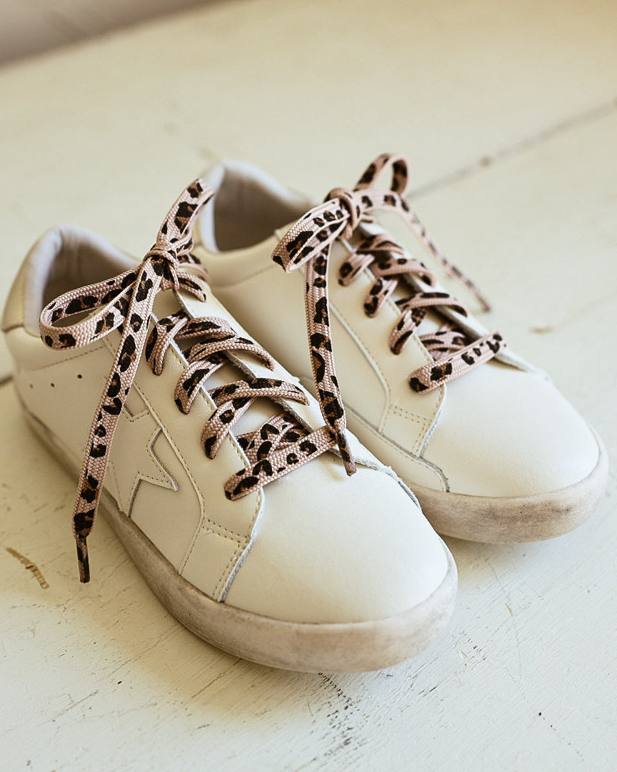 A pair of off-white leather low-top sneakers with tonal star cutouts on the sides, displayed on a rustic white wooden surface against a neutral beige wall. Leopard laces