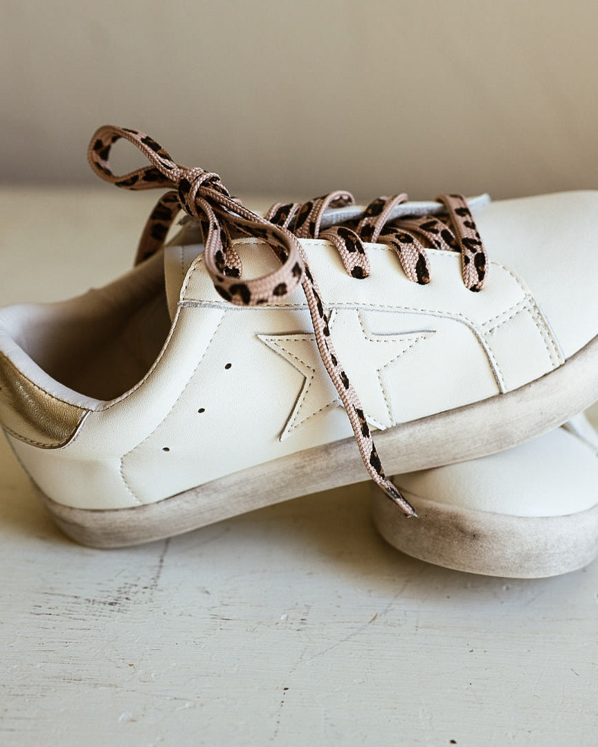 A pair of off-white leather sneakers with distressed detailing and leopard-print laces, resting on a light-colored wooden floor against a neutral beige background.