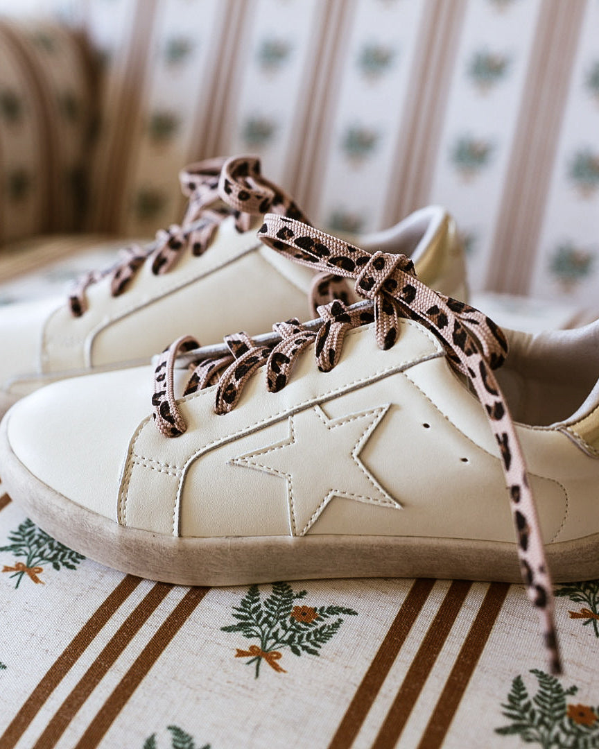 A pair of off-white leather sneakers with distressed detailing and leopard-print laces, resting on a light-colored wooden floor against a neutral beige background.