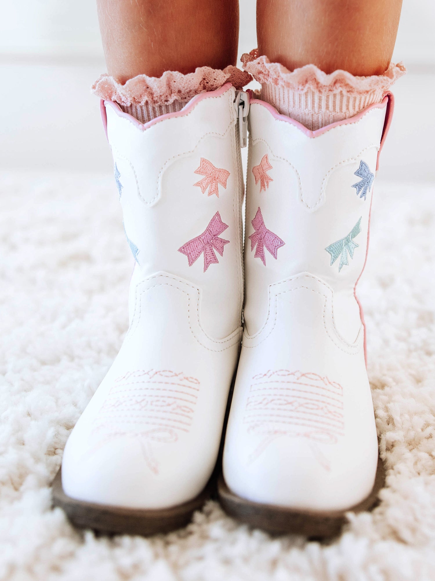 Step into sweetness with our Girls Bow Paneled Cowboy Boots, designed for twirls, giggles, and all-day play. These white faux-leather boots feature pastel pink, blue, and lavender star embroidery, charming bow panels, and an easy side zipper closure for quick on-and-off. With a soft interior lining and sturdy sole, these girls’ Western boots perfectly blend comfort and style—ideal for everyday wear, special occasions, or matching with her favorite SweetHoney dress.