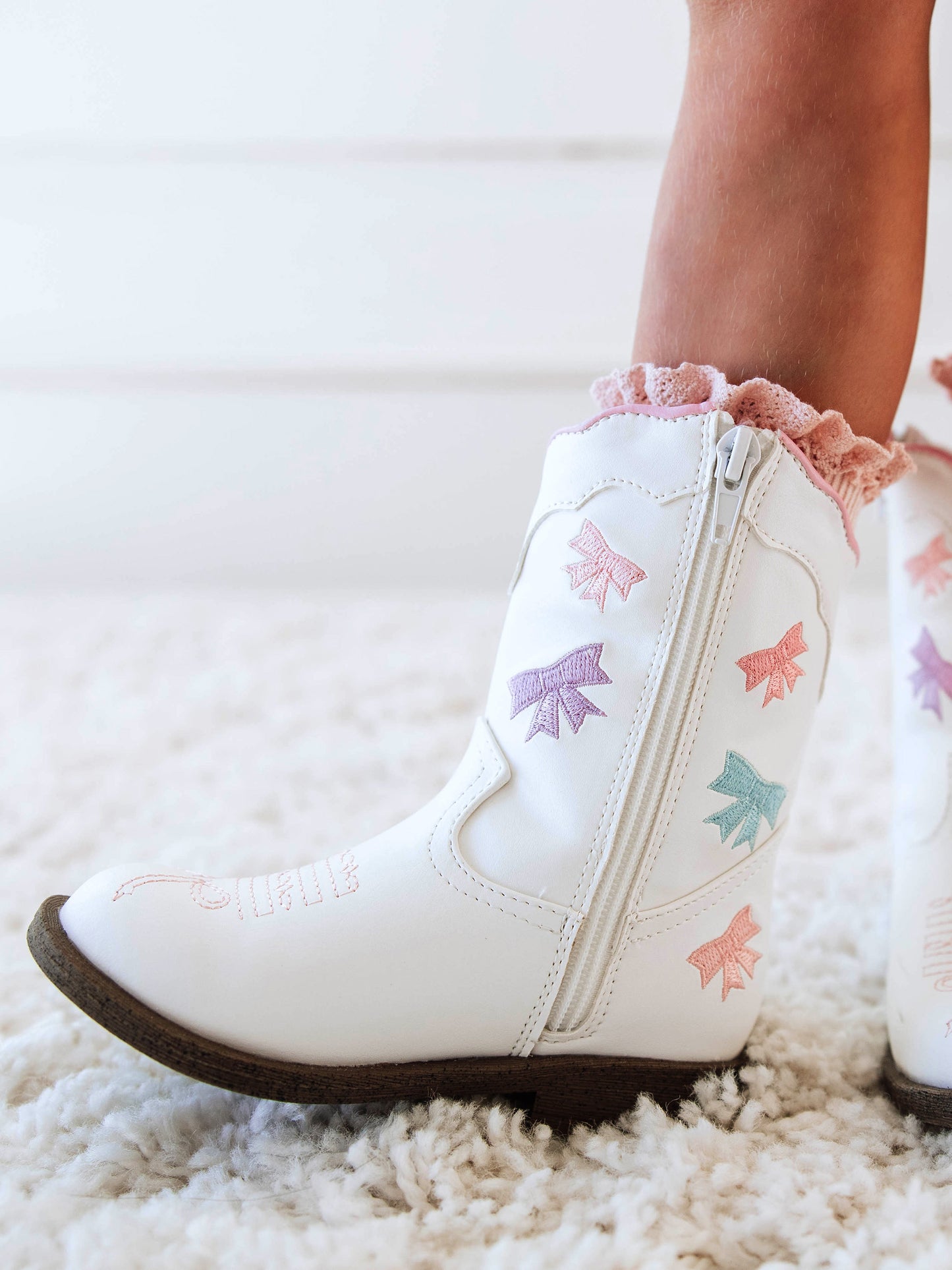 Step into sweetness with our Girls Bow Paneled Cowboy Boots, designed for twirls, giggles, and all-day play. These white faux-leather boots feature pastel pink, blue, and lavender star embroidery, charming bow panels, and an easy side zipper closure for quick on-and-off. With a soft interior lining and sturdy sole, these girls’ Western boots perfectly blend comfort and style—ideal for everyday wear, special occasions, or matching with her favorite SweetHoney dress.