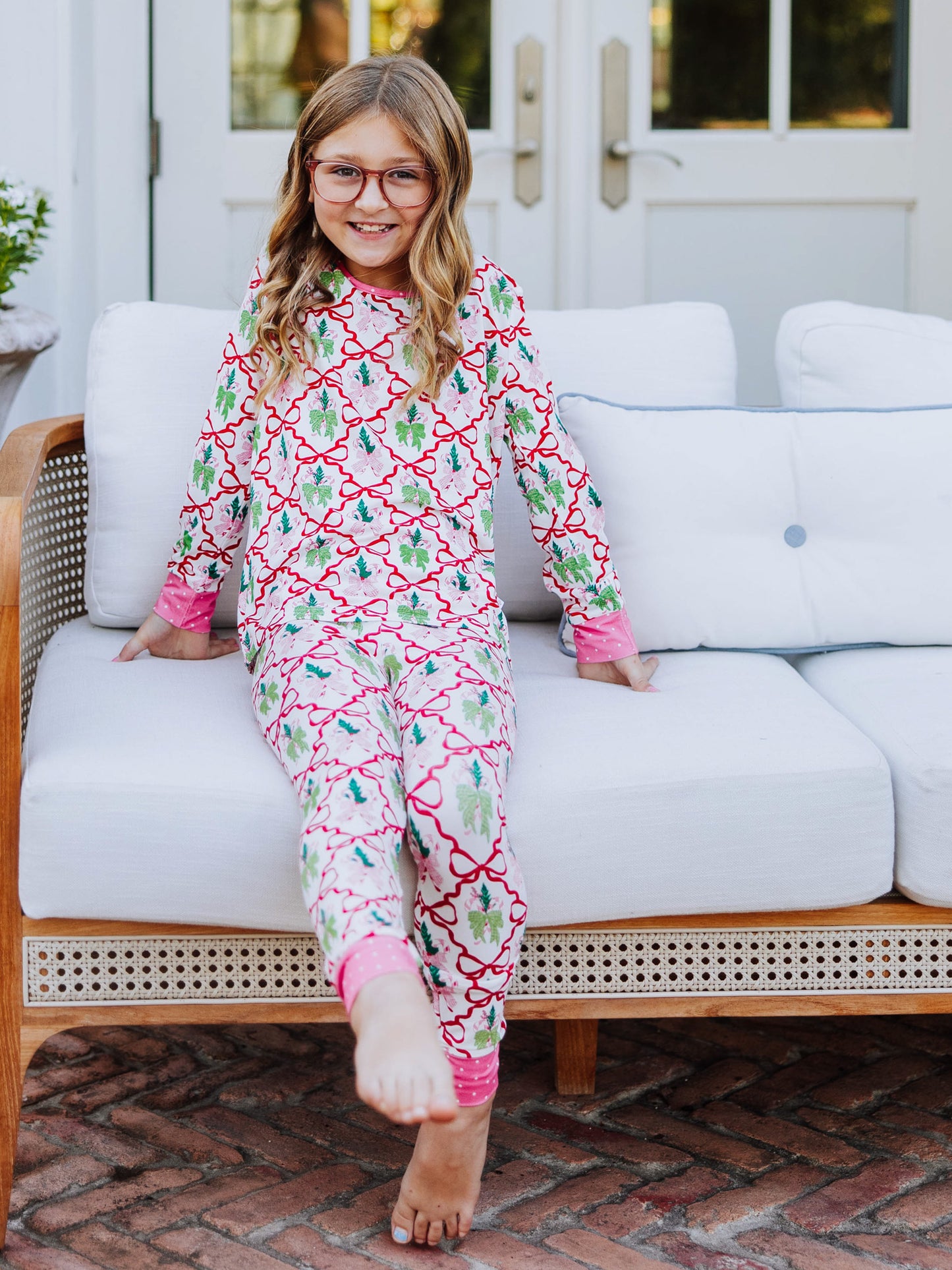 Girl walking outside on brick with couch in background. Model is wearing a matching two-piece set of pajamas that have colorful bows all over it. Model has leg out and it pointing toe.
