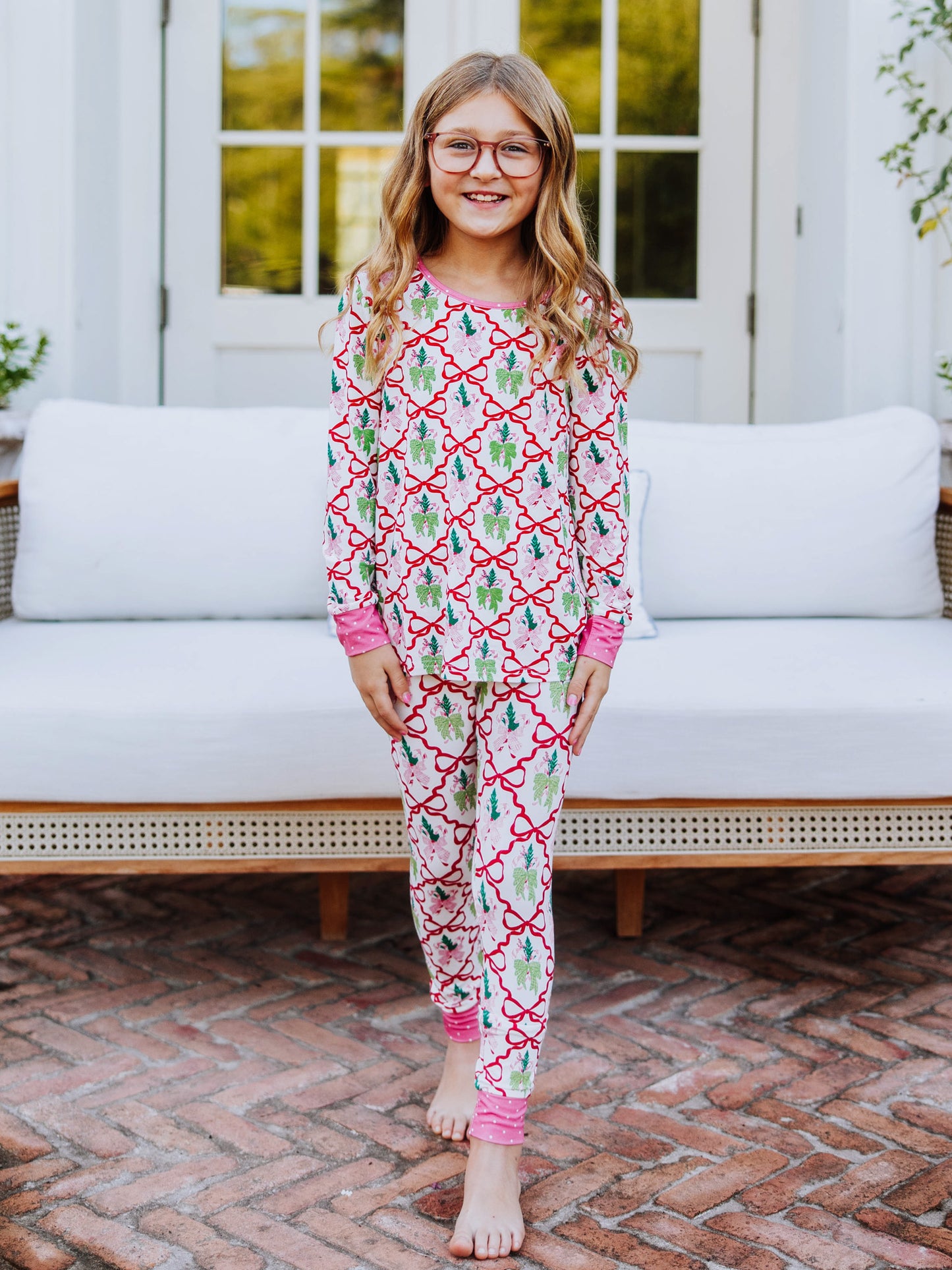 Girl walking outside on brick with couch in background. Model is wearing a matching two-piece set of pajamas that have colorful bows all over it. 