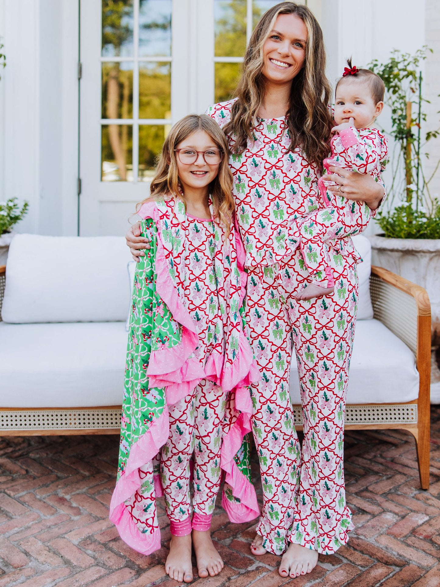 Family of three all wearing the same matching pajamas. Girl on left has matching blanket wrapped around her. The print is colorful and has red bows throughout. 