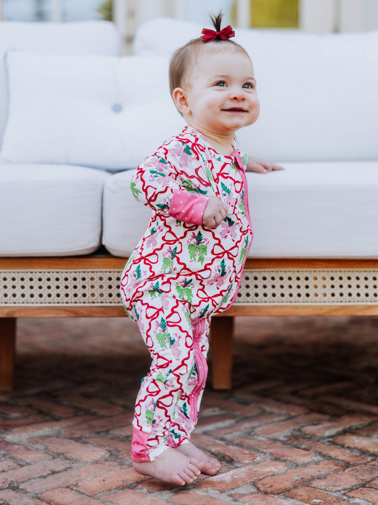 Baby standing up holding on to white couch smiling. Baby is wearing a colorful printed layette. 
