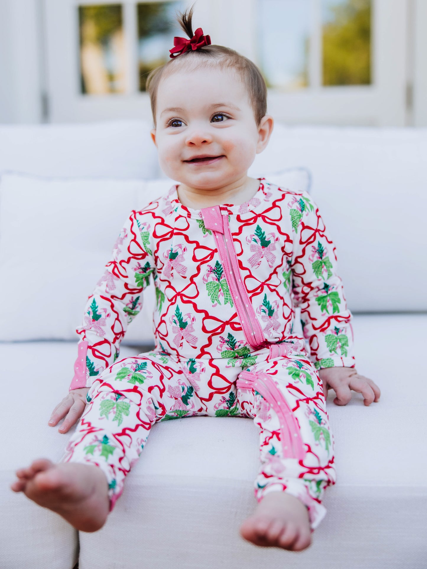 baby sitting on white couch smiling wearing a colorful layette with red bows.