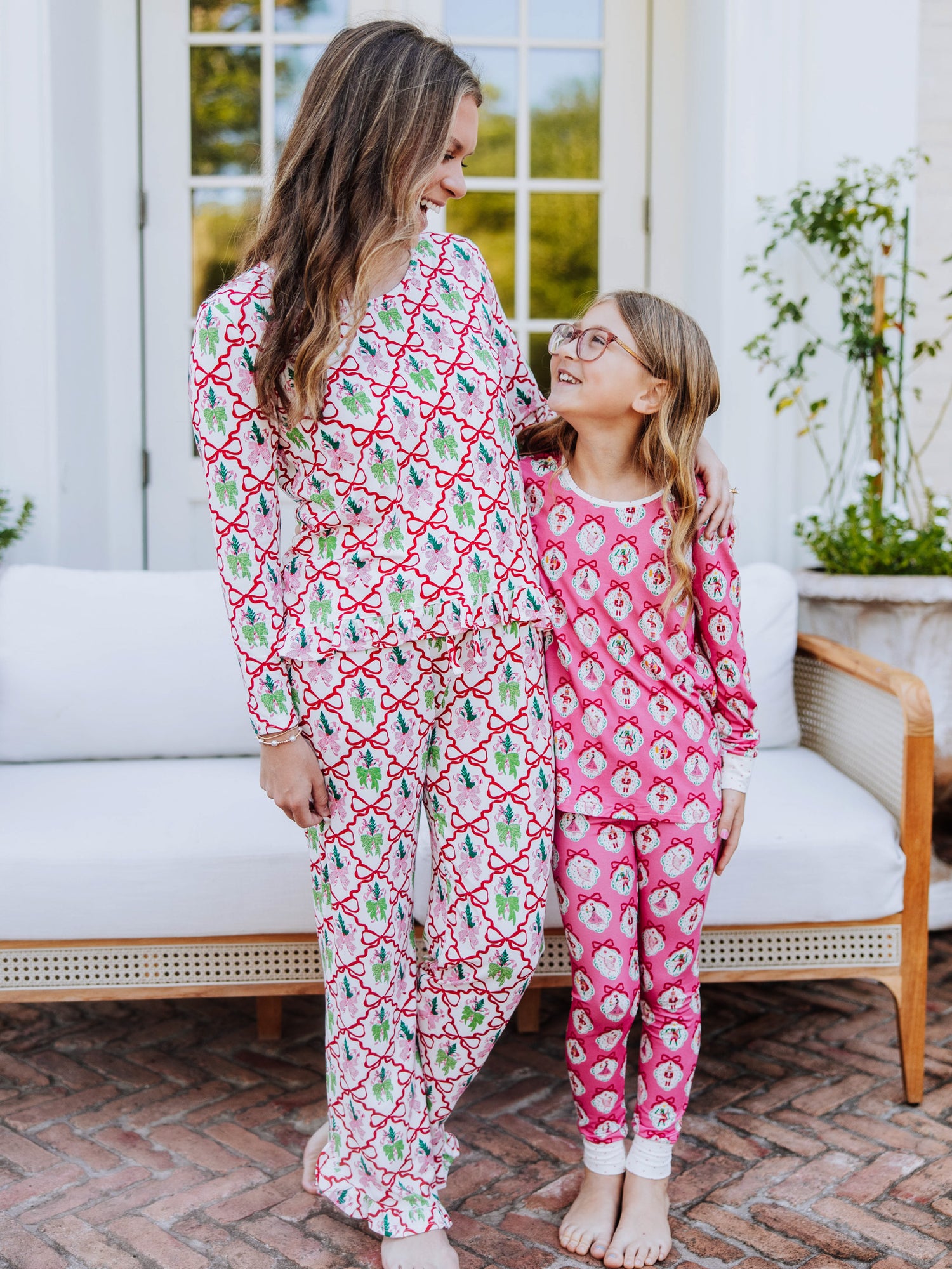 Women and girl standing outside in front of a white couch. women is wearing a colorful pair of christmas pajamas with bows on them and girl is also wearing a matching pair of fitted pajamas that have pink nutcrackers on them. Both are looking at one another. 