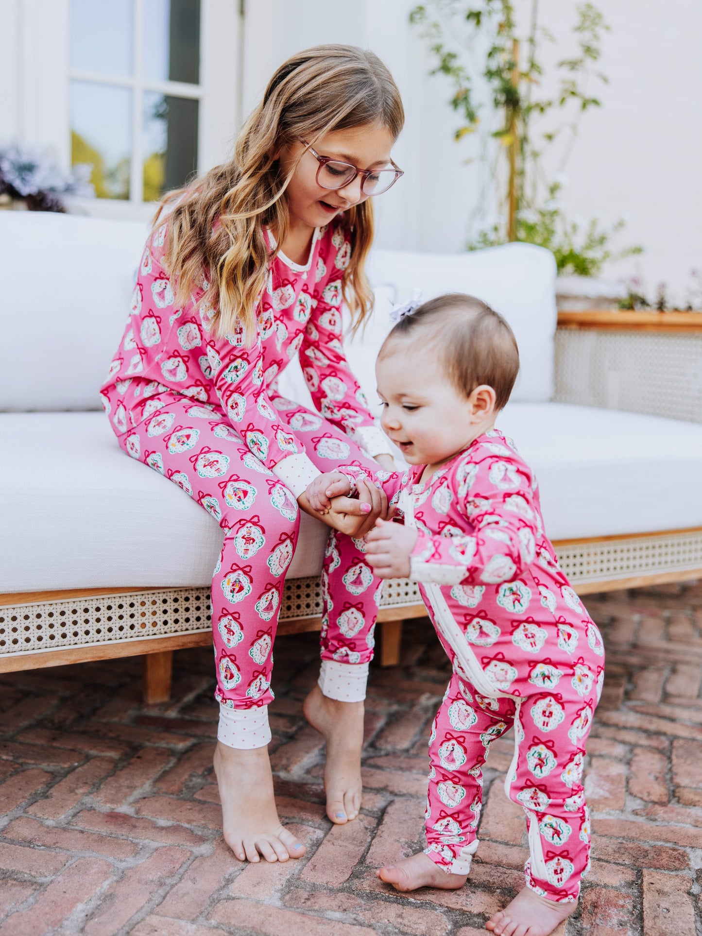 Girl holding on to babies hand. Both are wearing pajamas with matching pink nutcracker prints. 