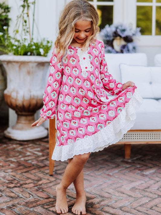 Girl standing outside on brick wearing a pink nutcracker printed dress. Model has feet crossed and is holding up the corner of one side of the dress. 