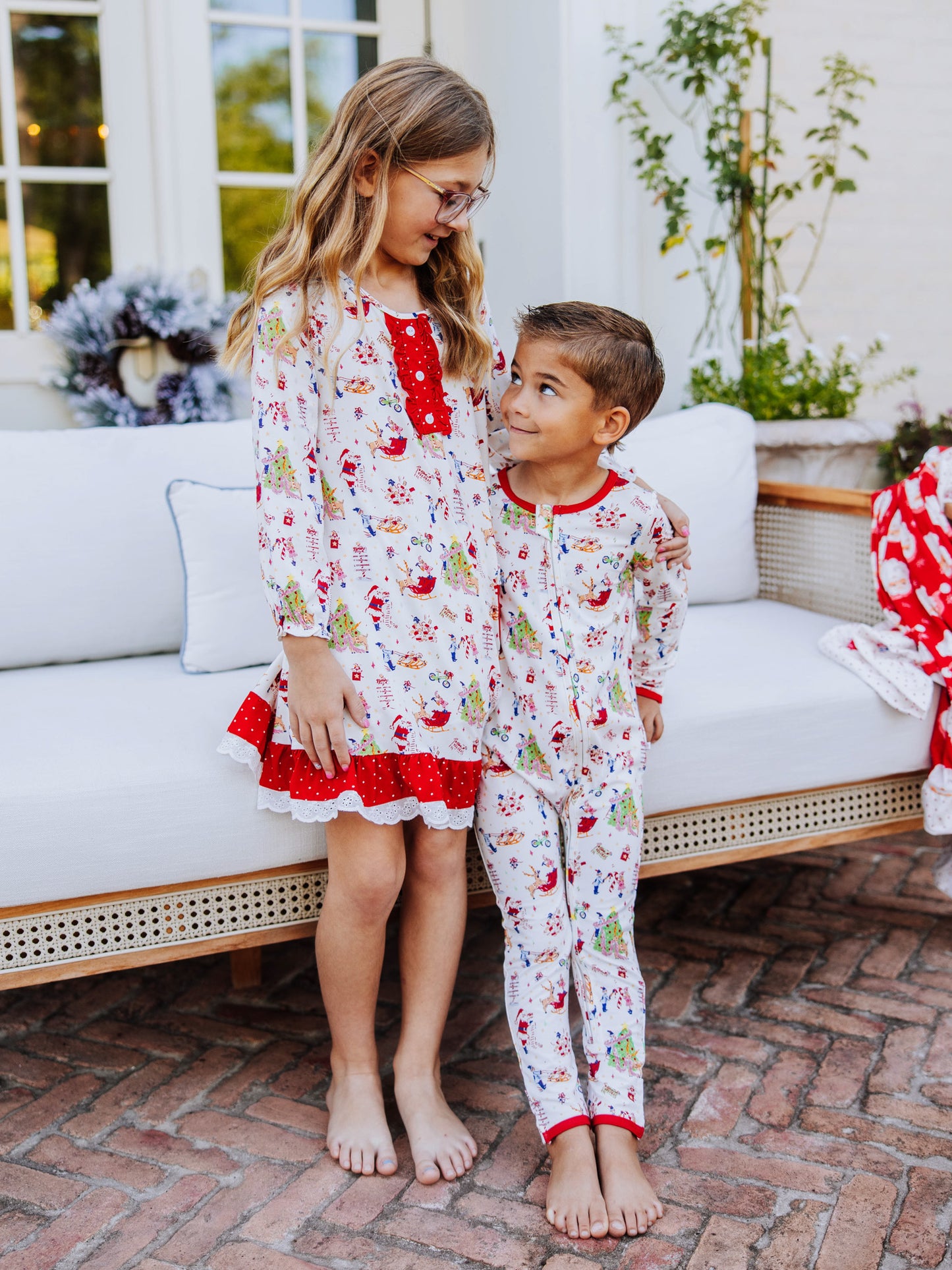 Girl and boy looking at one another standing outside against a white couch. Both are wearing the same north pole printed items. Girl is wearing a play dress and boy is in a layette.