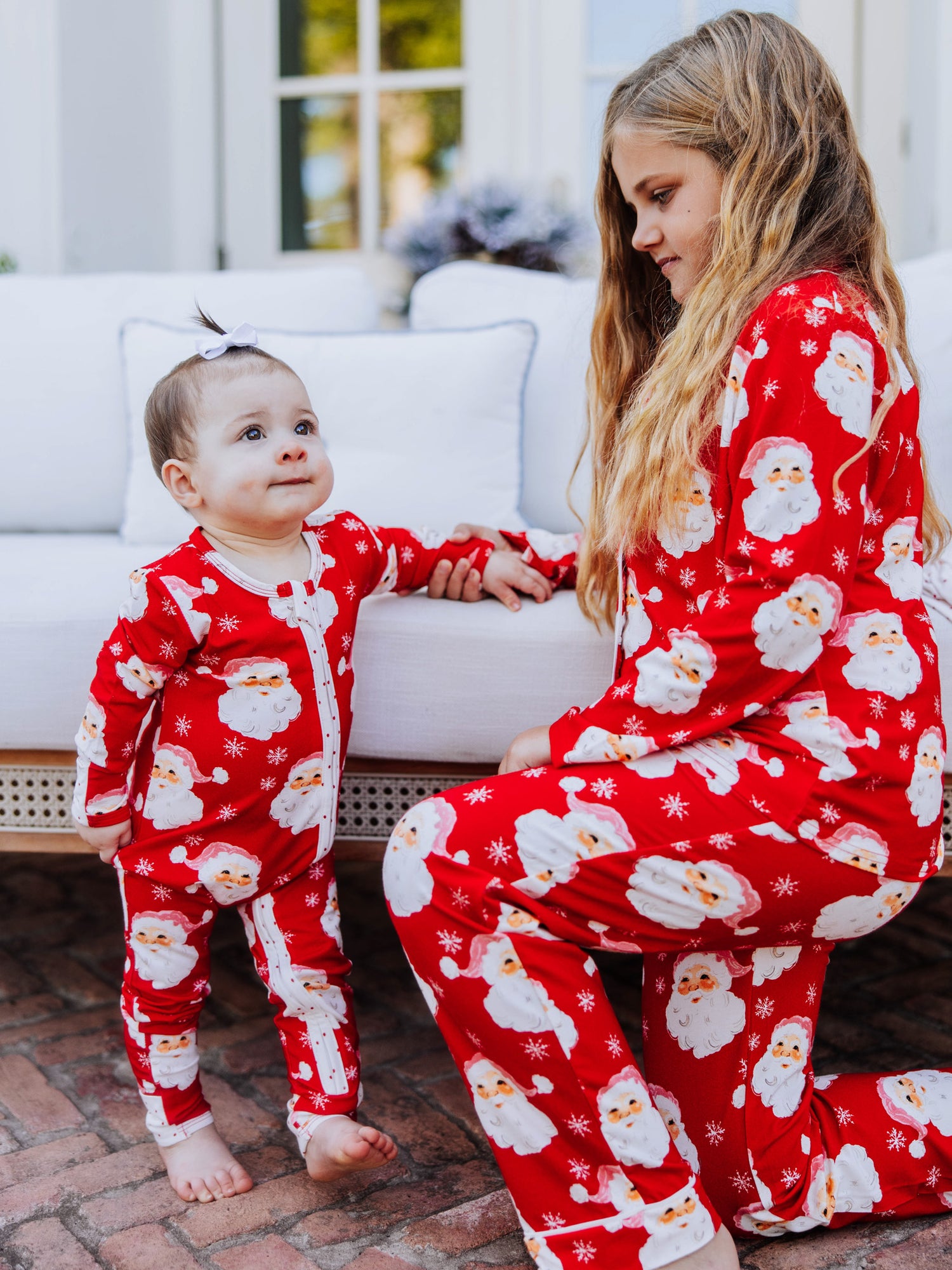 Baby girl standing up holding on to the side of a white couch. Baby is wearing a red santa layette. Kneelig beside her is an older girl wearing a two piece set in same print. 