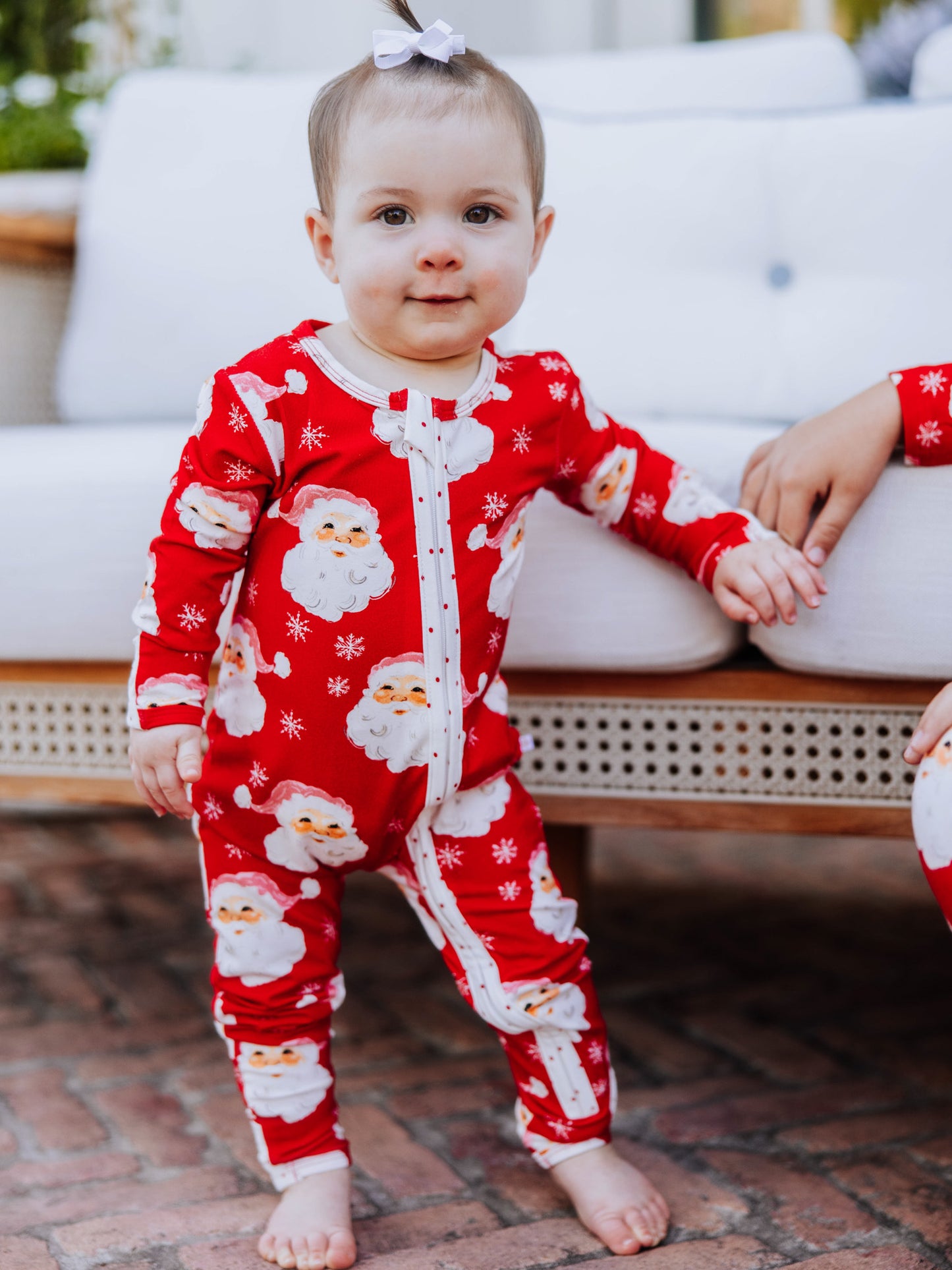 Baby girl standing up holding on to the side of a white couch. Baby is wearing a red santa layette. 