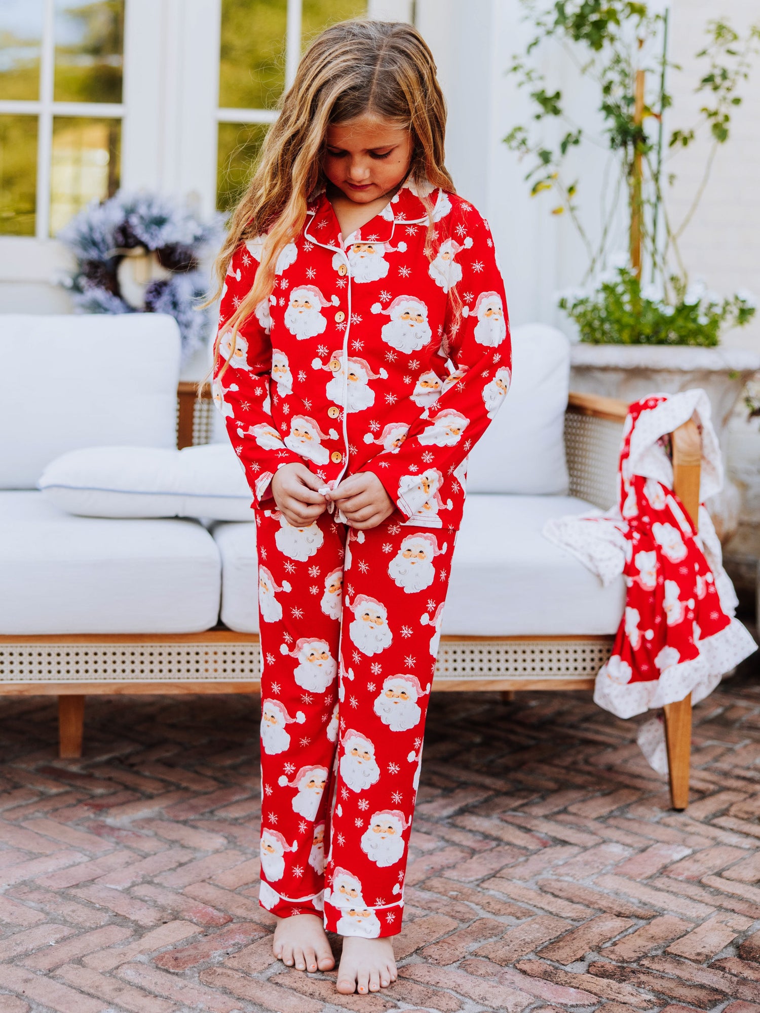 Girl standing on porch in front of a white couch and is wearing Santa pajamas with her hands in her pockets.