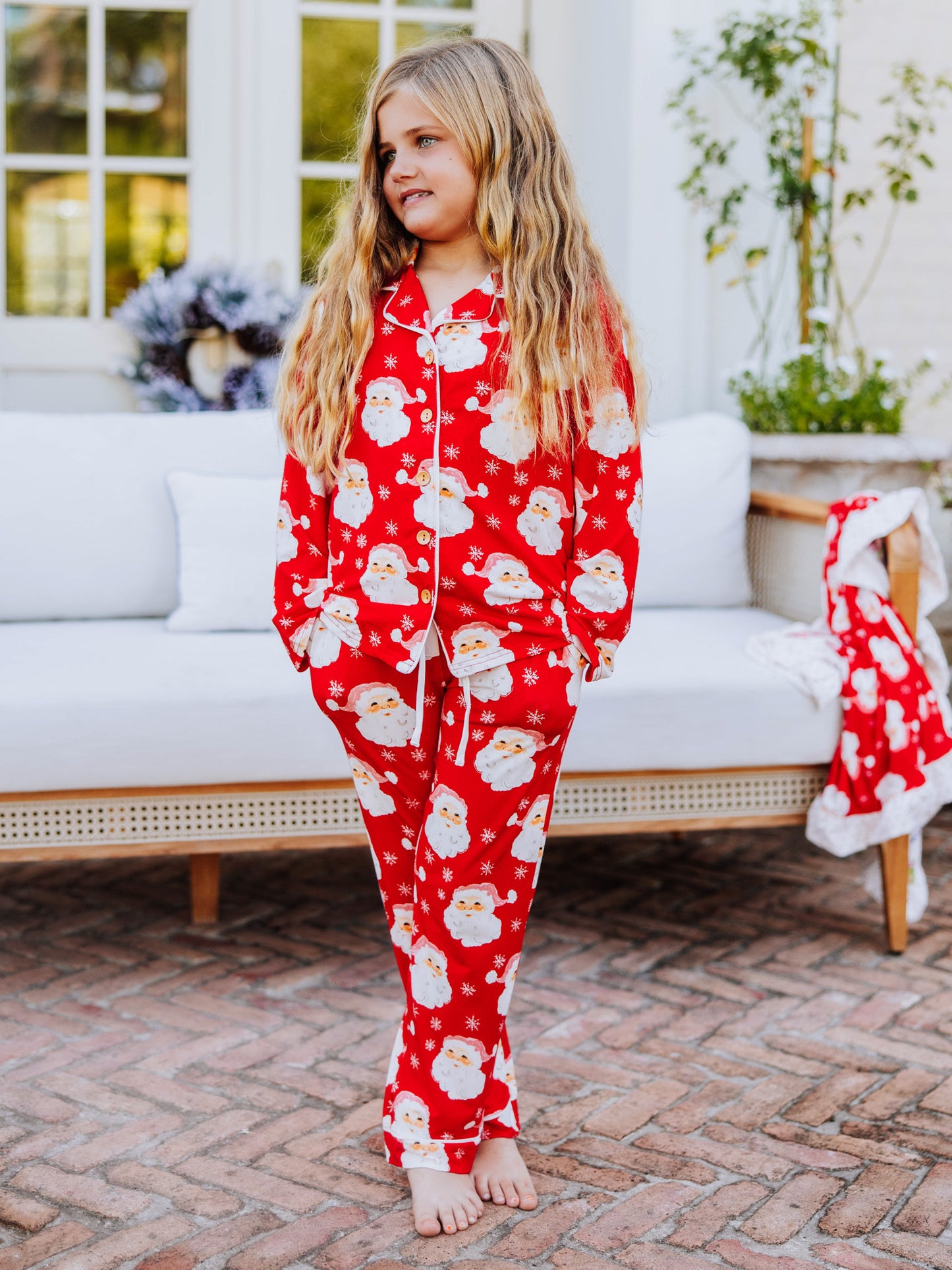 Girl standing on porch in front of a white couch and is wearing Santa pajamas with her hands in her pockets.